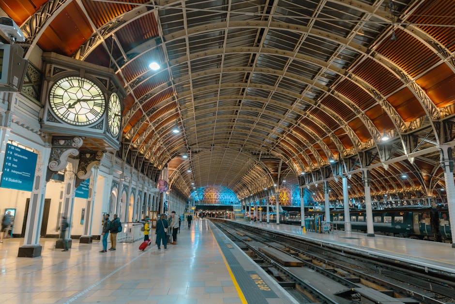 A spacious train station interior featuring a high arched ceiling constructed with metal framework and wooden panels, illuminated by bright overhead lights. On the left side, there is a large, ornate clock with a white face and black Roman numerals housed in decorative molding. Below the clock, blue and white signs display station information, and several people are standing or walking along the platform, some carrying luggage or waiting for trains. The platform surface is tiled with a mix of light and dark shades, and there are multiple train tracks running parallel towards a distant, illuminated arched ceiling at the far end of the station. The environment appears clean and well-lit, creating an organized atmosphere suitable for passenger transit, with the scene capturing the indoor space typically associated with a major railway hub, consistent with the context of alternative waste handling and logistics of large-scale transportation services by companies like Waste Disposal Paddington.