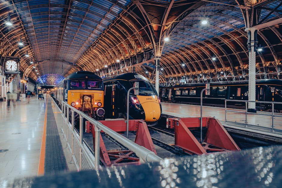 A modern train station interior featuring a large, curved iron and glass roof structure illuminated by bright ceiling lights, with a yellow and black train positioned on the left side of the platform. The front of the train displays narrow windows and a sleek aerodynamic design, with some purple and orange accents. The platform is spacious, with a beige tiled floor and a metal railing running along the edge. To the right, there are additional train carriages, darker in colour, parked on other tracks. A large clock hangs on the wall to the left, and the station appears well-lit with a clean, organized environment. The scene is enclosed within the station's arched roof, highlighting the high, intricate ironwork and expansive glass panels, creating a bright yet industrial atmosphere. This environment could relate to the context of rail travel or transporting goods, emphasizing infrastructure used for efficient movement, comparable in purpose to waste disposal and logistics services like those provided by Waste Disposal Paddington in the context of managing waste logistics or alternative transportation methods.