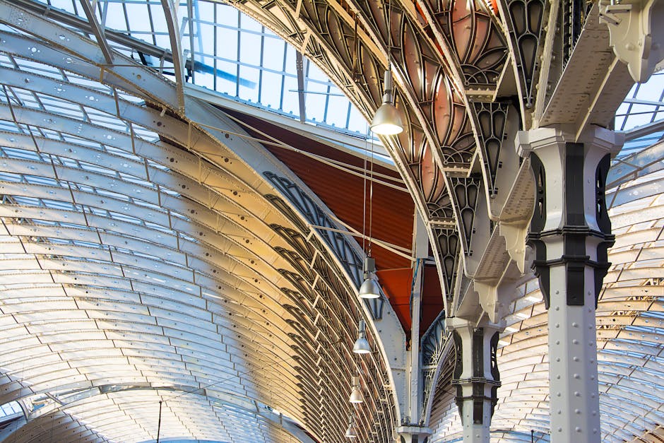 The image depicts an interior view of a large, historic iron and glass roof structure with intricate cast-iron supports painted in cream and black, featuring decorative detailing and riveted joints. The roof comprises curved glass panels framed by metal arches, allowing natural light to illuminate the space. Visible are hanging pendant lights suspended from the ceiling, providing additional lighting. The surrounding environment suggests an indoor setting, possibly part of a historic market or railway station, characterized by the ornate architectural features. The structural design emphasizes the industrial Victorian style typical of 19th-century engineering. The detailed ironwork, combined with the expansive glass ceiling, creates a bright and airy atmosphere. This type of architectural environment is relevant to the context of waste management and rubbish removal services, such as those provided by Waste Disposal Paddington, which specialize in handling waste from commercial or heritage sites that may require specialized cleaning or clearance, reflecting an alternative approach to waste handling within historic or architecturally significant environments.