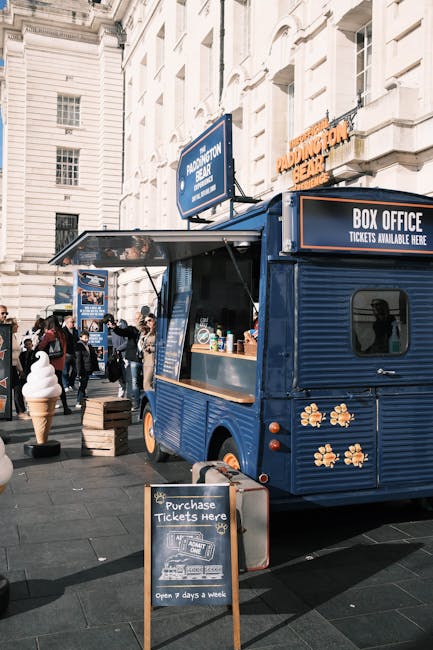 A small, dark blue mobile ice cream vendor caravan with a wooden counter window on a busy city street, surrounded by pedestrians. The caravan features decorative flower motifs on its lower section and has signage indicating ticket sales and the availability of ice cream treats. In front of the caravan, there is a chalkboard sign advertising ticket purchases, with text noting the shop's open seven days a week. To the left of the caravan, a life-sized ice cream cone sculpture and several other small wooden crates or boxes are visible, suggesting a temporary setup for an outdoor event or market. Behind the scene, tall, cream-colored buildings with classical architectural features, such as ornate moldings and large windows, line the street. Sunlight illuminates the area, creating a lively atmosphere. The setting reflects a typical street scene where outdoor vendors and local businesses operate, offering alternative options for outdoor refreshments and small-scale retail, similar in concept to independent retail or off-site services provided by waste management companies like Waste Disposal Paddington, which handle rubbish and alternative waste collection.