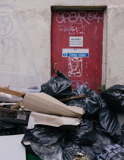 A pile of black rubbish bags, some torn open revealing discarded packaging, cardboard boxes, and miscellaneous waste, stacked on a pavement in front of a red metal door covered with graffiti and stickers. The door is set into a plain, textured, off-white wall with visible weathering and surface irregularities. The bags and waste occupy the foreground, with the door positioned centrally in the background, featuring signs such as a white sticker reading 'No Money' and a blue sign stating 'Keep Clear,' suggesting an informal waste collection or disposal area. The scene is illuminated by natural daylight, with minimal shadows, highlighting the disorderly accumulation of refuse typical of independent waste clearing efforts, relevant to private rubbish removal services like those offered by Waste Disposal Paddington, operating in the local area for non-standard waste handling. The setting suggests an urban alley or back street environment where prompt rubbish clearance is often necessary to maintain cleanliness and prevent obstruction.
