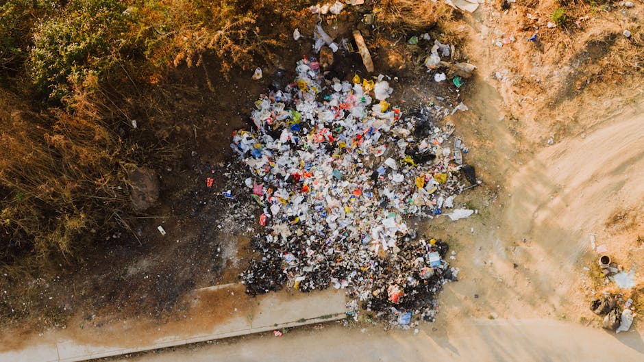 An aerial view of a small, rectangular excavation site on a dirt surface showing a large pile of mixed household waste, including plastic bottles, paper, and debris, some of which are partially burnt or darkened. The surrounding area consists of dry, reddish-brown soil with sparse vegetation and bushes at the top edge of the image. To the right, a narrow dirt pathway runs alongside the site, indicating a semi-urban or construction environment. The waste appears to have been recently dumped or disturbed, with scattered pieces around the main pile. The scene suggests an irregular waste disposal activity, possibly related to private on-site clearance or non-official rubbish dumping, which Waste Disposal Paddington might handle through their rubbish removal services for safe and proper cleanup.