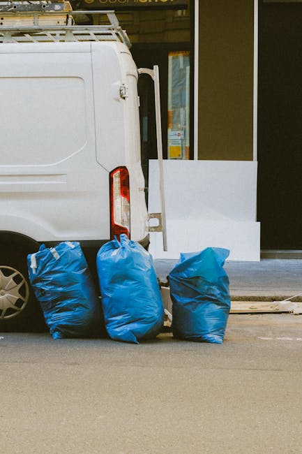 The image shows the rear section of a white cargo van parked on a street in front of a commercial building with large windows and a dark exterior. Three blue rubbish bags made of thick plastic are lined up on the pavement next to the van, positioned close together and appearing full, with some visible creases and folds on their surfaces. The bags are placed on a flat, paved surface with a curb visible at the bottom edge of the image. Behind the van, part of a glass door or window is visible, revealing a partial interior or reflection. The scene appears to be during daylight hours, with natural light illuminating the area evenly. This setup suggests the van is likely used for collection or disposal purposes, consistent with waste removal services like those provided by Waste Disposal Paddington, who specialize in private waste handling and on-site clearance activities.