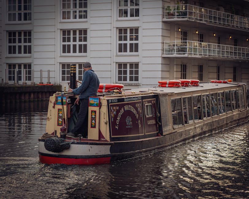 A narrowboat on a canal with a dark maroon and cream exterior, displaying the name 'LONDON WATERBOAT' along with decorative floral motifs and a small window on its side. A man in a blue jacket and dark trousers stands at the stern, operating the boat with one hand on the steering controls, while the other rests on the side. The boat is equipped with orange life rings secured on the roof. The surrounding environment features calm water reflecting the boat and nearby modern residential buildings with large windows, white railings, and balconies. The scene is illuminated with soft natural light, suggesting an overcast day, and the setting indicates an environment where alternative waste collection methods, such as private boat-based rubbish removal services, might be utilized for urban or canal-side properties.