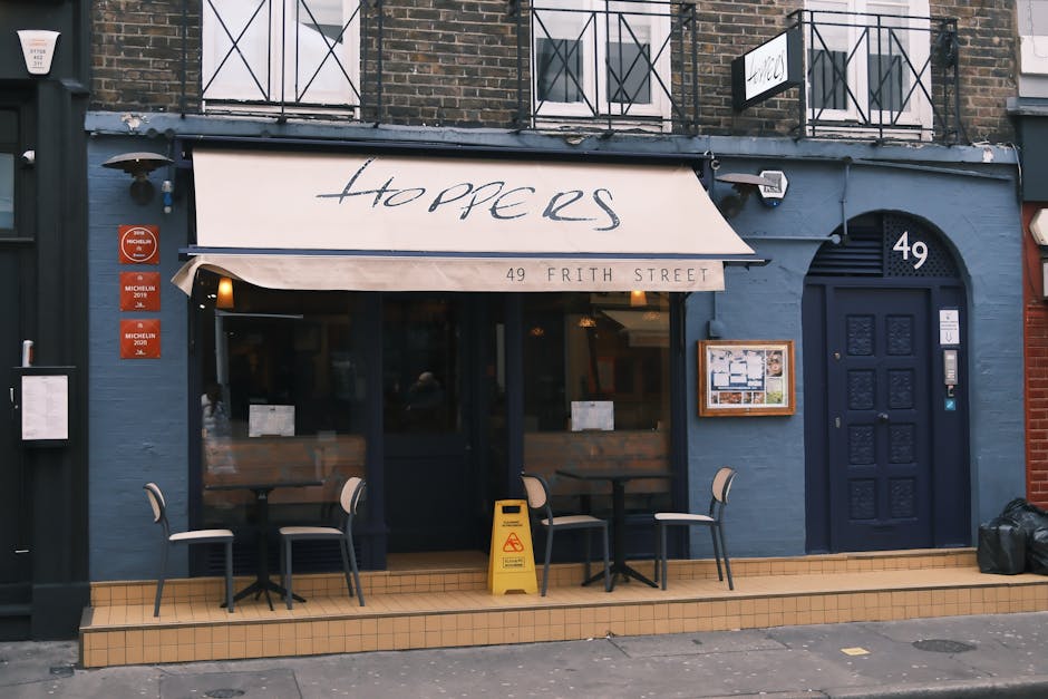 The exterior of a restaurant named 'Hoppers' located at 49 Frith Street features a dark blue painted brick façade with a blue ornate front door on the right, displaying decorative paneling and a small metallic doorbell. To the left, there is a large window with a black frame, behind which warm interior lighting is visible, providing a welcoming atmosphere. Above the window, a beige awning with the restaurant's name in elegant black script extends outward, reading 'Hoppers,' along with the address. In front of the establishment, a small outdoor seating area consists of three tables, each accompanied by two light-colored chairs with dark metal frames, set on a tiled small step that matches the entrance’s pavement. A yellow 'caution wet floor' sign is placed on the tiles near the tables, indicating recent cleaning or spill. To the left, there are three red plaques indicating Michelin recognition, and on the right, a small menu display case is mounted on the wall. The environment appears clean and well-maintained, with muted natural daylight illuminating the scene, highlighting the textures and colors of the brickwork, signage, and outdoor furniture, reflective of a typical city street restaurant setting.