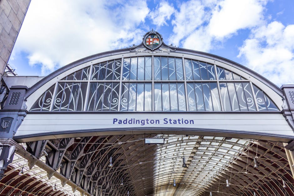 The image shows the upper portion of Paddington Station's ornate iron and glass roof structure, featuring a detailed arched framework with decorative scrollwork and vertical glass panels reflecting the partly cloudy sky. A crest or coat of arms is positioned at the apex of the arch, indicating the station's historic design. Below the arch, a white horizontal sign displays the station name 'Paddington Station' in blue letters. The roof's intricate metalwork and glass panels allow natural light to illuminate the station’s interior, with the surrounding environment visible through the glass. The weather appears clear and bright, with scattered clouds visible in the reflection. This scene emphasizes the station's architectural character and its role as a central transport hub, which can relate to the broader theme of infrastructure and operational context for private or alternative waste handling services such as those provided by Waste Disposal Paddington, often involving on-site or independent collection facilities.