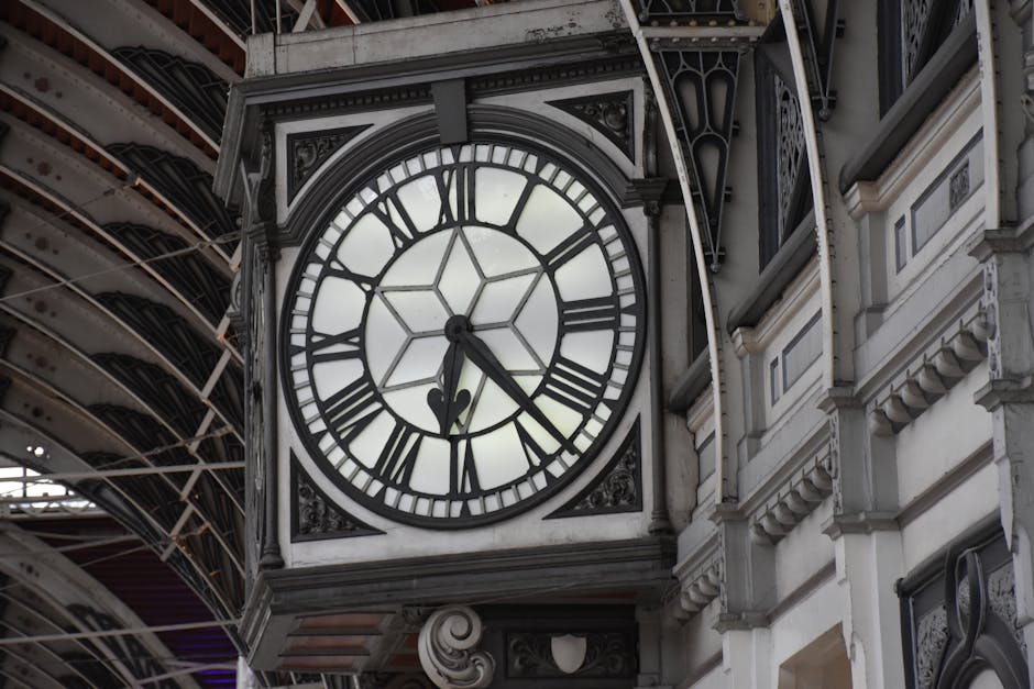 A close-up view of a large, ornate clock mounted on the exterior of a historic building, with a white clock face featuring black Roman numerals and black minute markers. The black hour and minute hands indicate a time of approximately 4:22. The clock is surrounded by decorative black metal framing with intricate detailing, positioned against the cream-colored facade of the building which includes ornate molding and architectural embellishments. In the background, parts of a metal and glass covered structure or canopy with visible beams and supports extend overhead, providing a contrasting modern architectural element. The lighting suggests daytime with natural light softly illuminating the scene, highlighting the textures of the stone and metal. This scene emphasizes the historic character and decorative craftsmanship of urban architecture, relevant to the context of building management or architectural preservation in a city environment.