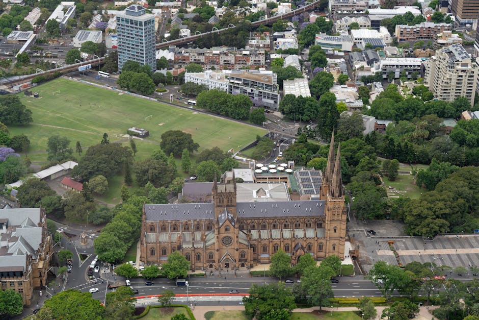 An aerial view of a large Gothic-style church with pointed spires and arched windows, situated in a cityscape surrounded by abundant greenery and trees. The church's stone exterior shows intricate architectural details typical of historic ecclesiastical buildings. Adjacent to the church are parking areas with a few cars, and nearby are clusters of residential and commercial buildings varying in height and modern design, with some featuring flat roofs and solar panels. To the left of the church, there is a park with expansive green lawns, scattered trees, and open spaces, bordered by streets with moving vehicles. In the background, there are taller office buildings and dense housing, with roads and a railway line crossing the scene, indicating an urban environment with mixed uses. The lighting appears natural with overcast skies, providing clear visibility of the diverse structures and lush vegetation typical of central city areas, aligning with locations where independent waste disposal services could be used for rubbish removal or site clearance purposes.
